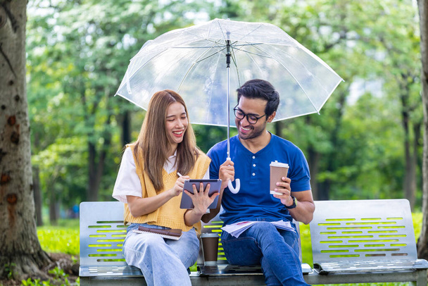 Νεαρό ασιατικό και ινδικό ζευγάρι απολαμβάνουν στιγμές χαλάρωσης στη βροχή μαζί στο δημόσιο πάρκο, ενώ κάθονται μαζί στον πάγκο κατά τη διάρκεια του Σαββατοκύριακου - Φωτογραφία, εικόνα
