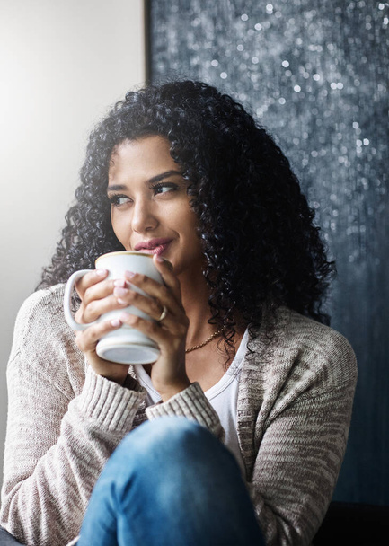 Ik heb geen zin om naar buiten te gaan. een vrolijke jonge vrouw genietend van een kop koffie terwijl ze overdag op een stoel thuis zit - Foto, afbeelding