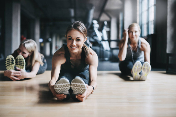 Κάθε άσκηση ξεκινά με ζέσταμα. Πλήρες πλάνο τριών ελκυστικών και αθλητικών γυναικών που εκτείνονται πριν από την προπόνηση τους στο γυμναστήριο - Φωτογραφία, εικόνα