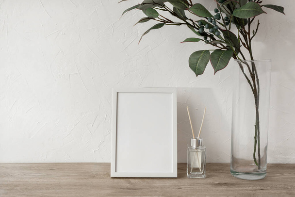 Blank photo frame, green leaves on vase, home fragrance on a beige wooden tabletop, white plaster wall background, minimalist brand or interior design template, mockup with copy space - Φωτογραφία, εικόνα