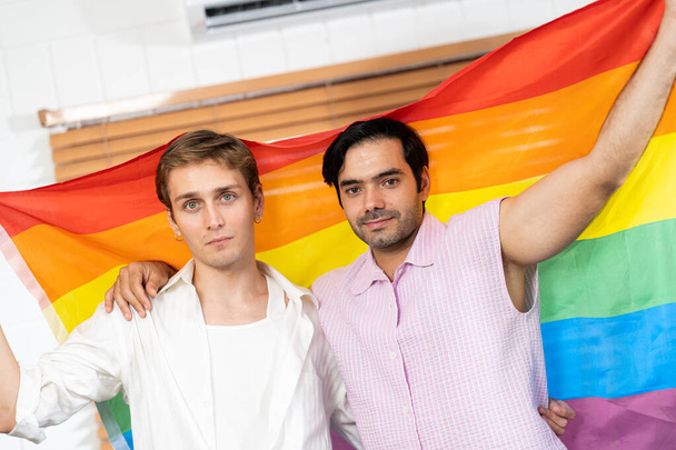 gay paar holding lgbt regenboog vlag binnen thuis. selectieve focus. gay paar omarmen en tonen hun liefde met regenboog vlag. LGBT en liefde concept. gay trots en regenboog achtergrond. - Foto, afbeelding
