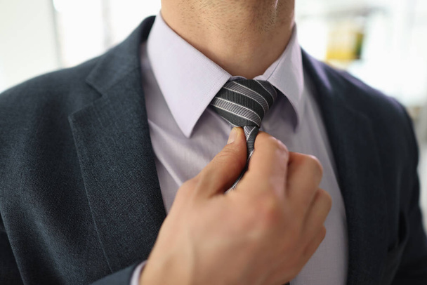 Successful businessman adjusts striped tie with hand to feel comfortable. Young man in classic formal suit feels stressed at work in office - Фото, зображення