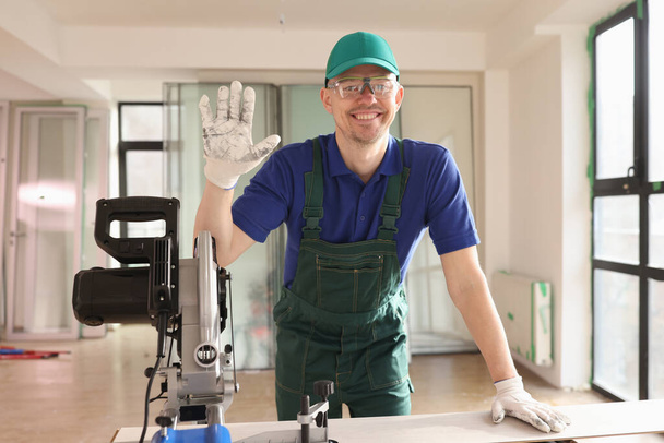 Cheerful foreman in uniform shows hello looking in camera and smiling. Carpenter processes laminate with machinery equipment for flooring - Photo, Image