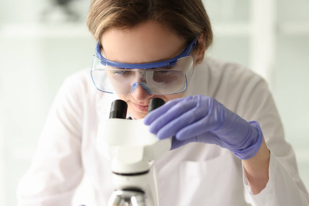Koruyucu gözlüklü kadın bilim adamı elektronik mikroskop kullanarak örnek inceler. Laboratuvar kadın çalışanı bilimsel araştırma yapıyor. - Fotoğraf, Görsel
