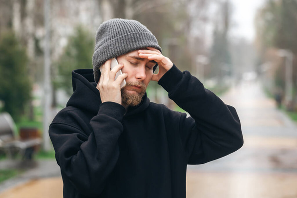 Brodaty mężczyzna w kapeluszu rozmawia przez telefon w parku i trzyma głowę, złe wieści, problemy i trudne pytania. - Zdjęcie, obraz