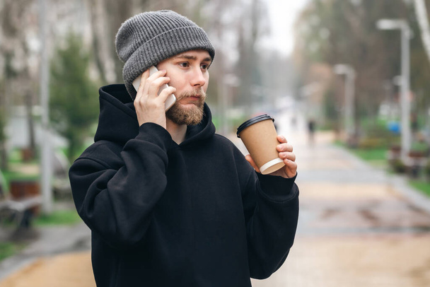 Jeune homme avec une barbe et un chapeau parlant au téléphone et buvant du café dans une tasse en papier dans le parc, fond flou. - Photo, image