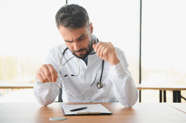 Handsome doctor man wearing medical uniform sitting on his workplace tired holding his head feeling fatigue and headache. Stress and frustration concept. - Foto, imagen