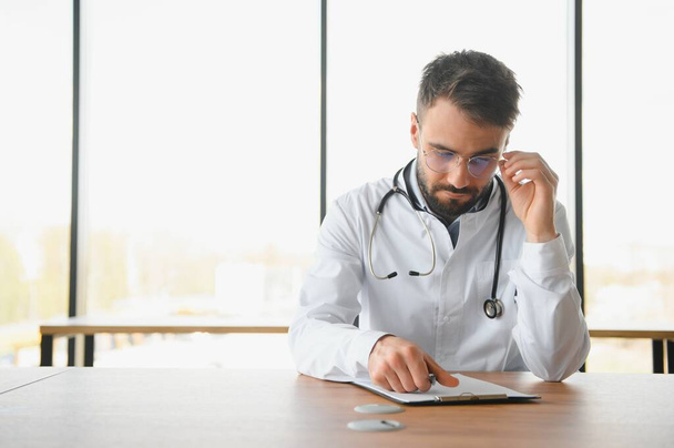 Handsome doctor man wearing medical uniform sitting on his workplace tired holding his head feeling fatigue and headache. Stress and frustration concept. - 写真・画像
