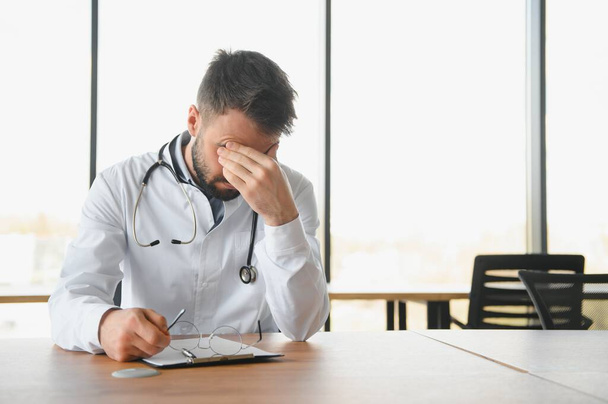 Handsome doctor man wearing medical uniform sitting on his workplace tired holding his head feeling fatigue and headache. Stress and frustration concept. - Foto, afbeelding