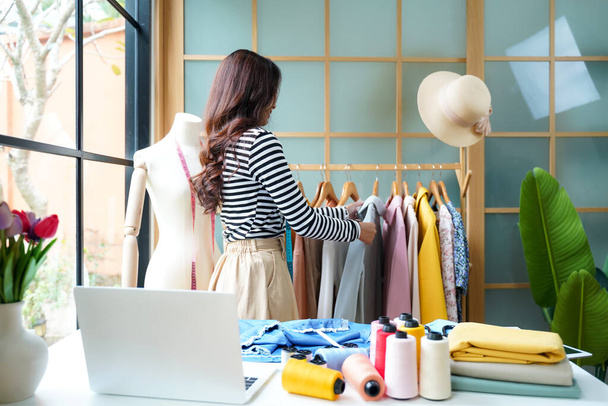 Young women fashion designer using laptop working and design tailor and cloth in the showroom,  Lifestyle Stylish tailor taking measurements on mannequin for cutting cloths in studio.  Business and Industry small Concept  - 写真・画像