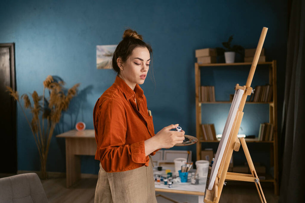 young brazilian woman artist in home studio poses stands near canvas opens tube of paint. Black hair, dressed in casual clothes. concept of creativity and hobby. copy space. - Foto, imagen