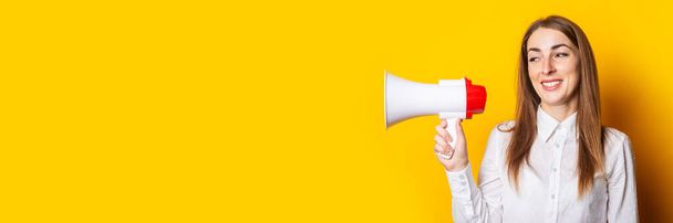 Friendly young woman holds a megaphone in her hands on a yellow background. Hiring concept, help wanted. Banner. - Fotó, kép