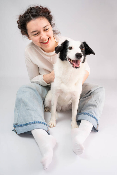 Dog and woman smiling sitting on gray background. Owner young girl in blue jeans and white jumper cuddling with her cute pet border collie young dog. Positive happy emotions. Wide open smile - Photo, image