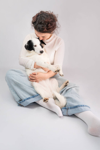 Dog owner and dog cuddling. Hugging young woman and adorable white border collie pet sitting on the floor. grey (gray) background. Casual blue jeans and white clothes. Best friends trust relationship - Photo, image