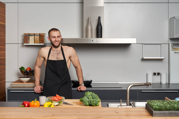 Happy shirtless muscular man wearing a chef apron coocking a salad in modern kitchen. - 写真・画像
