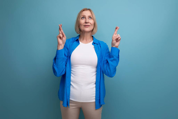 femme mature aux cheveux blonds en chemise décontractée doigts croisés et pense sur fond bleu avec espace de copie. - Photo, image