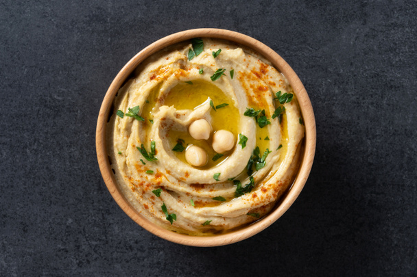 Chickpea hummus in a wooden bowl garnished with parsley, paprika and olive oil on black slate background. Top view - Fotografie, Obrázek