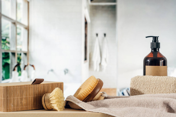 Kolekce péče o tělo v koupelně se sprchovým gelem v lahvičce na dávkovač, masážní bambusové kartáče, čistý měkký ručník, koupací houba a proutěný koš na dřevěné polici. Ekologické produkty pro každodenní hygienu - Fotografie, Obrázek