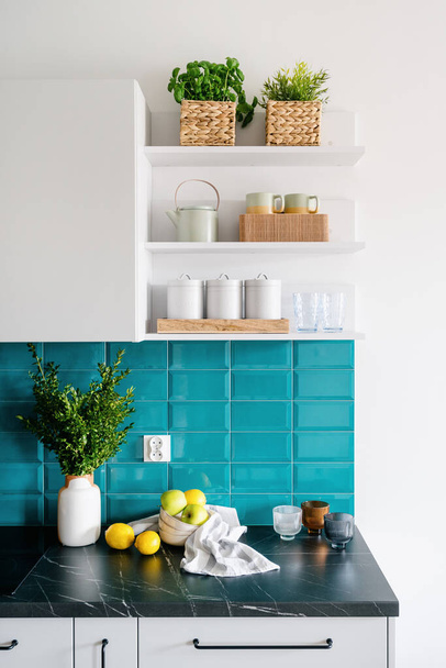 stylish interior at home kitchen with white furniture cupboard, dark marble countertop and blue tile on wall, wicker basket with plants, ceramic storage jars, cups and glasses - Foto, Imagem