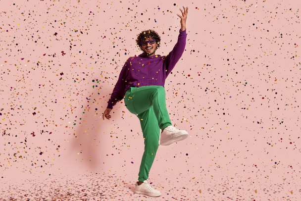 Full length of playful Indian man dancing and throwing confetti against pink background - Fotoğraf, Görsel
