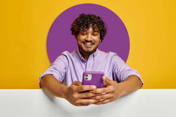 Handsome Indian man leaning on copy space and using phone against yellow background - 写真・画像