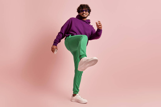 Playful young Indian man stretching out leg while dancing against pink background - Valokuva, kuva