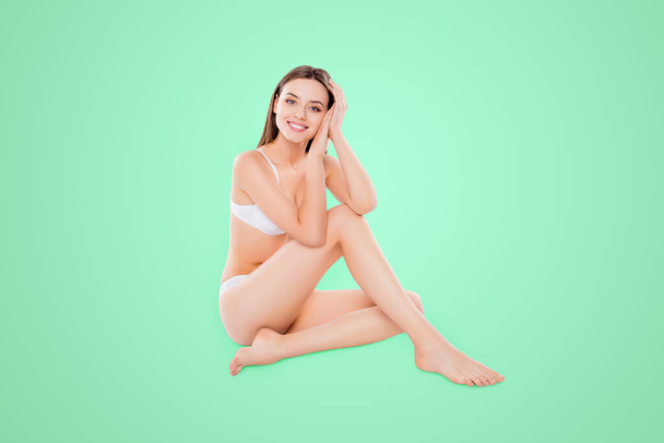 Portrait of thin slender woman sitting over white background in bra and bikini looking at camera demonstrating perfect body plain skin. - Foto, imagen