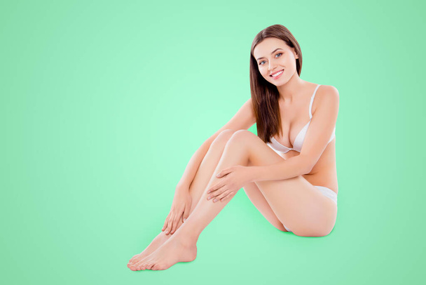 Portrait of slim thin woman sitting over white background in bra and bikini enjoying touching plain legs after depilation. - Foto, Imagen