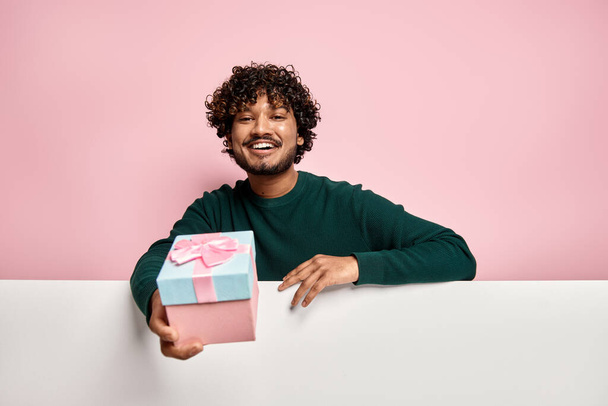 Happy Indian man stretching out gift box while congratulating you against pink background - Photo, image
