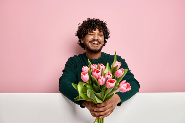 Handsome Indian man stretching out flower bouquet and winking against pink background - Photo, image