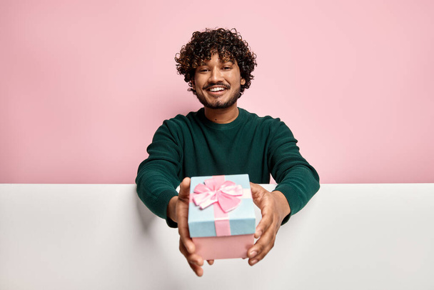 Handsome Indian man stretching out gift box against pink background - Photo, image
