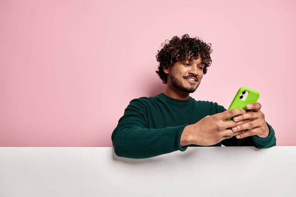 Handsome Indian man leaning on copy space and using smart phone against pink background - Fotó, kép