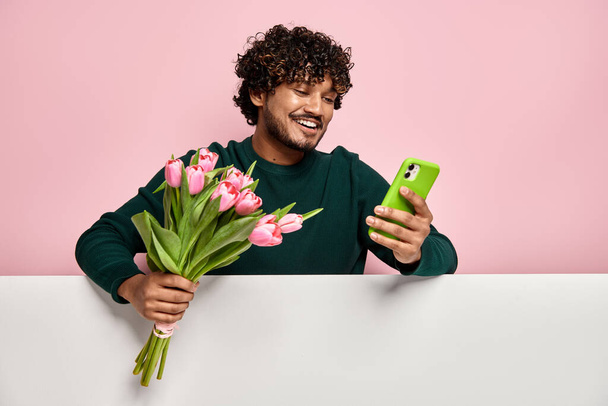 Handsome Indian man holding flower bouquet and using smart phone against pink background - Fotó, kép