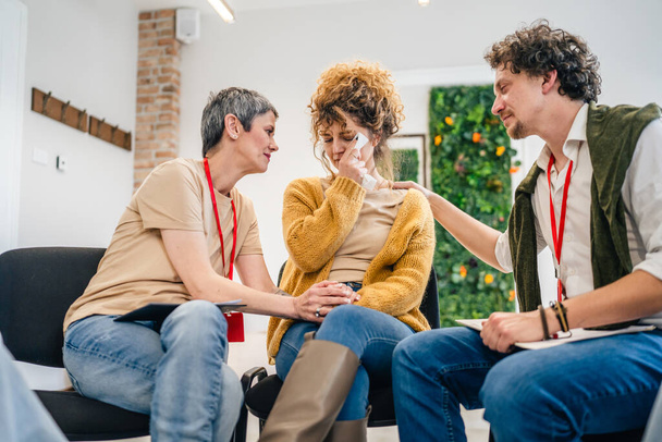 Un grupo de personas sentadas en un círculo con dos terapeutas y una mujer en el centro discutiendo y brindando apoyo emocional para ella. - Foto, imagen