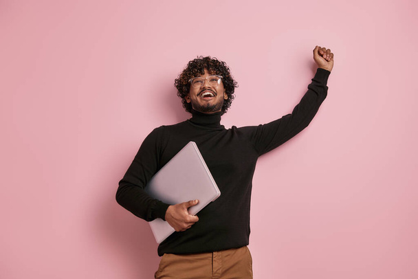 Joyful Indian man carrying laptop and gesturing against pink background - 写真・画像