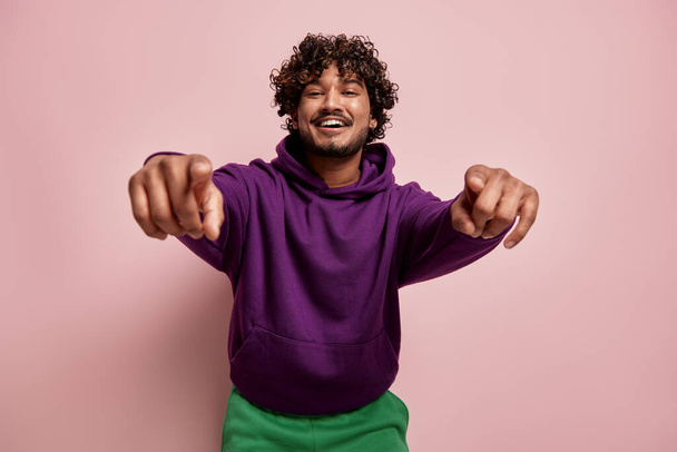 Happy young Indian man pointing camera and smiling against pink background - Photo, Image