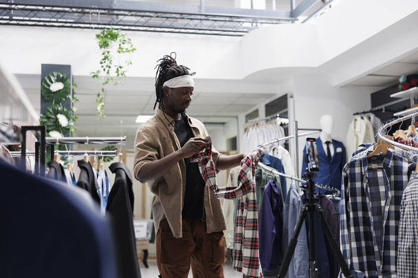 Influenceur afro-américain parlant de la marque de vêtements en magasin tout en enregistrant des vidéos sur smartphone. Blogueur de mode sur les médias sociaux montrant chemise et donnant avis pour les abonnés en ligne - Photo, image