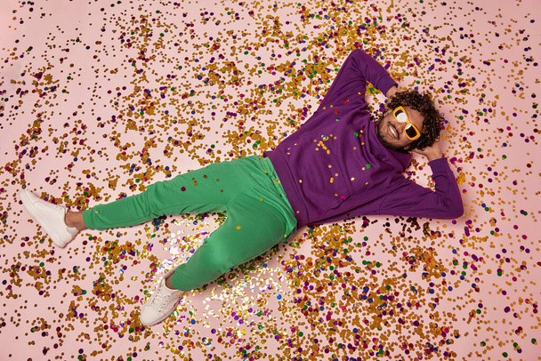 Relaxed Indian man holding hands behind head and smiling while lying on colorful confetti - Foto, imagen