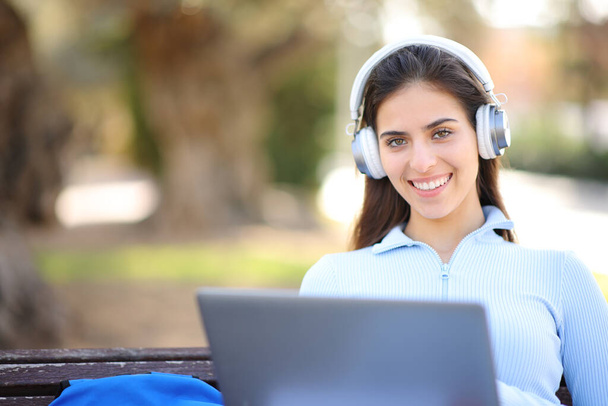 Happy student with laptop and headphone looks at you in a park - Fotoğraf, Görsel