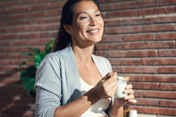 Знімок красивої веселої вагітної жінки з брекетами, що їдять йогурт на кухні вдома
 - Фото, зображення