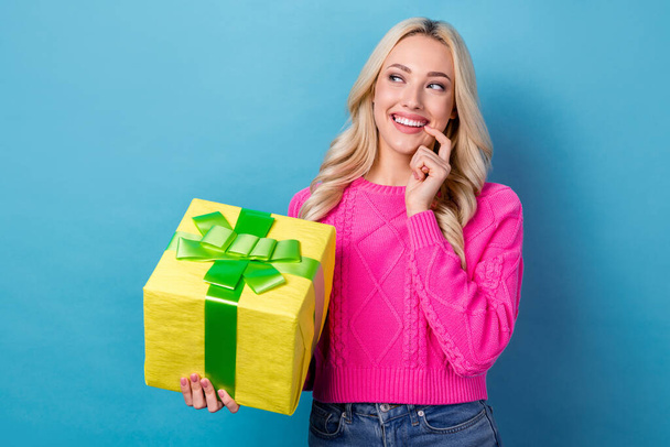 Photo of nice girl with curly hairdo knit sweater holding present box look empty space finger teeth isolated on blue color background. - Zdjęcie, obraz