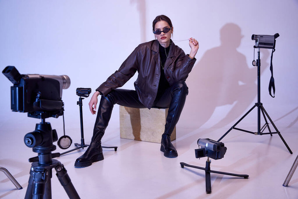 Brutal look. Beautiful young girl in stylish, trendy clothes, leather jacket, pants, sunglasses and accessories. Retro cameras in studio. Fashion, style, retro and vintage, gadgets, technology concept - Фото, изображение