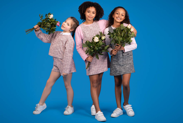 Boldog nemzetközi kislányok barátnők csokrok virágok élvezik tavasszal elszigetelt kék háttér, stúdió. Ünnepi ünneplés, gratuláció, gyermekkori és gyermekkori érzelmek - Fotó, kép
