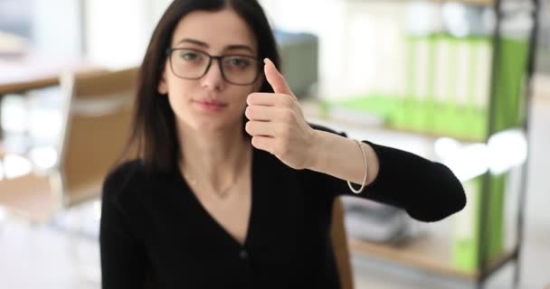 Mulher morena com óculos mostra aprovação sinal de polegar no local de trabalho. Administrador com sorriso se senta à mesa contra a premissa turva câmera lenta - Filmagem, Vídeo