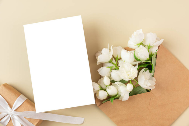 Blank greeting card with white jasmine flowers and gift box on beige background. Wedding invitation. Mock up. Flat lay. Mothers day layout - Photo, Image