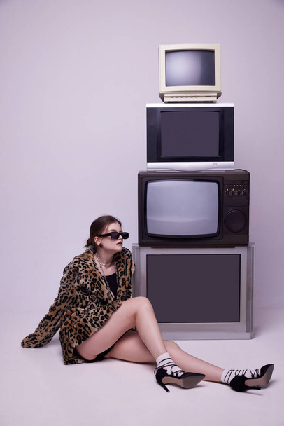 Influencer. Young girl in stylish animal print coat and trendy sunglasses posing near retro TV sets against grey studio background. Concept of fashion, 80s, 90s style, retro and vintage, mass media - 写真・画像