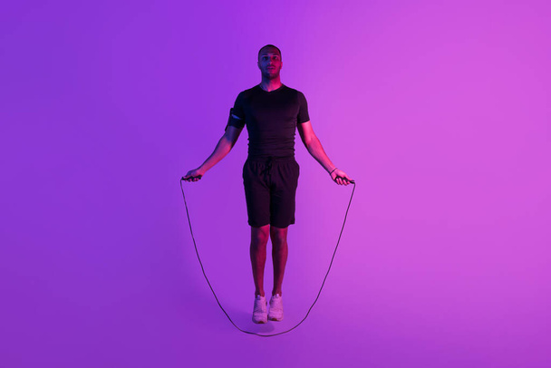 Zwarte sportman Oefenen met Jumping Rope Fitness Workout Poseren Over Purple Neon Studio Achtergrond. Volledige lengte schot van sportieve man training doen sprongen kijken naar camera - Foto, afbeelding