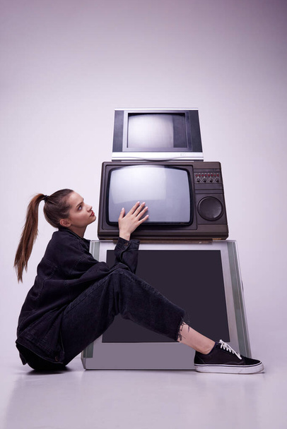 Addiction to mass media. Pretty young girl in black clothes sitting near retro TV sets against grey studio background. Fashion, 80s, 90s style, retro and vintage, gadgets, beauty, technology concept - Foto, afbeelding