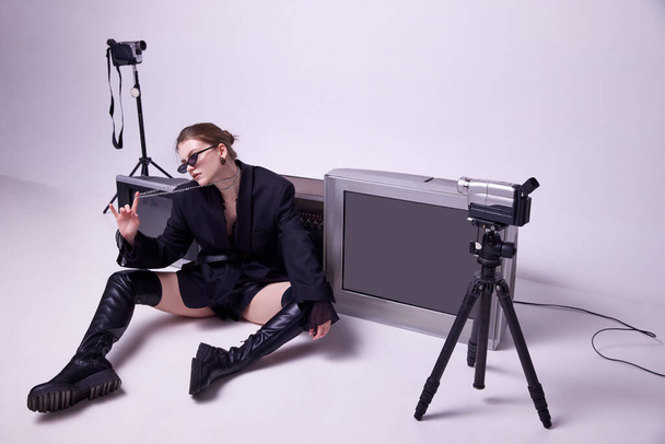 Young girl in stylish black jacket, leather boots and sunglasses sitting on floor around retro TV sets and camera. Concept of fashion, 80s, 90s style, retro and vintage, gadgets, beauty, technology - Photo, image