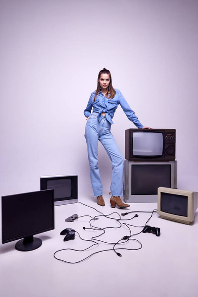 Young girl in jeans clothes posing around many retro TV sets and console. Game popularity, leisure activity. Concept of game, leisure activity, 80s, 90s style, retro and vintage, gadgets - 写真・画像